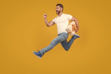 Energetic courier man running with gift box in midair yellow studio background. Fast courier delivery. Courier service.