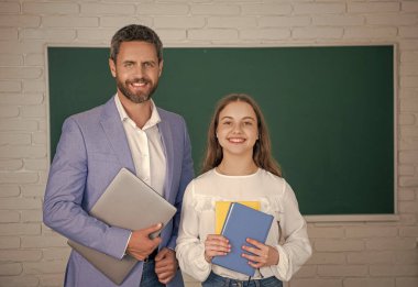 gülümseyen çocuk öğretmeniyle tahtada duruyor dizüstü bilgisayar ve çalışma kitabı.