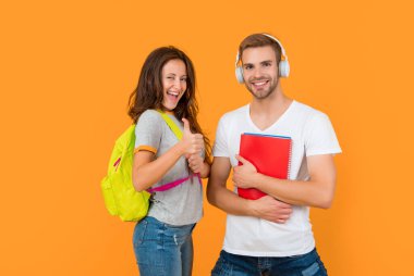 Sarı arka planda çalışma kitabı ve sırt çantası olan kulaklıklı neşeli iki öğrenci başparmağını kaldırdı ve bilgi sahibi oldu..