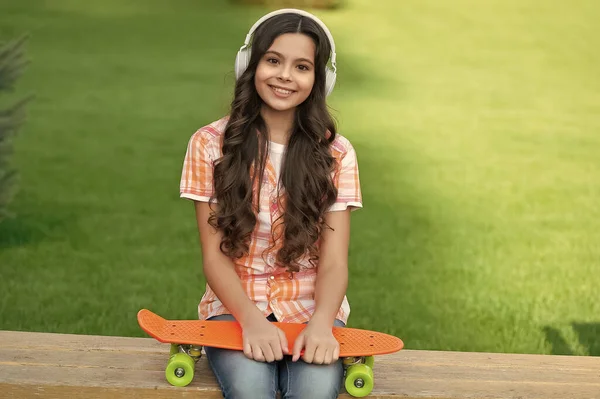 スケートボードを持つ肯定的な10代の女の子の写真は音楽を聞く スケートボードのアウトドアを持つ10代の女の子 ヘッドフォンでスケートボードを持つ10代の女の子 外にスケートボードを持つ10代の女の子 — ストック写真