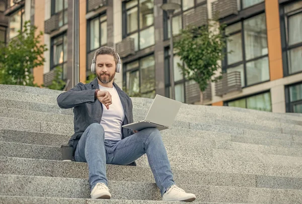 stock image photo of man check time for webinar online on laptop with headphones. man webinar online outdoor. man webinar online in the street. man webinar online outside.