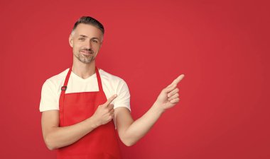 mature barista in apron on red background. point finger.