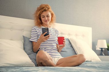 Telefonda internette kahve sörfü yapan gülümseyen bir kadın. Evde telefonuyla internette sörf yapan bir kadın. Kadının evinde internet sörfü.