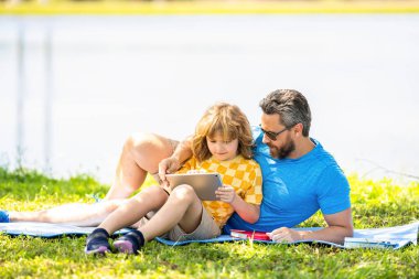Baba ve oğul parkta dinleniyor. çocukluk okulu eğitimi. Oğlu ve babası yazın kaynaşıyorlar. Baba ve oğul eğitimi almış. Baba oğul eğitimini kitap ve tabletle şekillendirir. yeşil doğa.