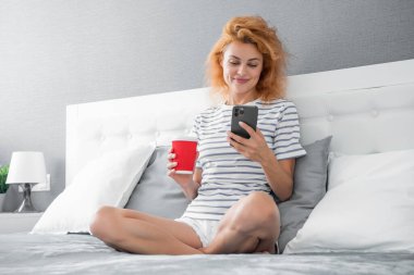 Telefonda internette kahve sörfü yapan gülümseyen bir kadın. Evde telefonuyla internette sörf yapan bir kadın. Kadının evinde internet sörfü.