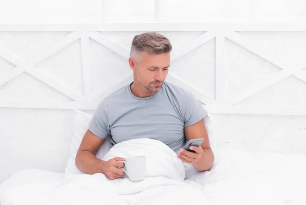 Mobile text messaging. Awake man send text message in bed. Messaging from smartphone. SMS texting. Cellular communication. Morning relaxation with cup of tea or coffee.