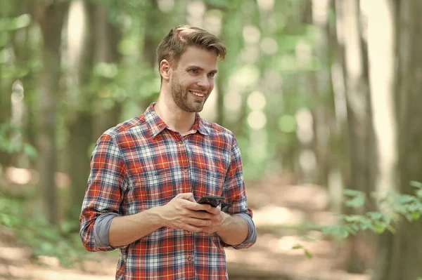 Adam dışarıda telefonda gülümsüyor ve mesajlaşıyor. Adam dışarıda telefonla mesajlaşıyor. Telefondaki mesajlaşan adam kareli gömlek giyiyor. Ormanda telefonla mesajlaşan bir adamın fotoğrafı..