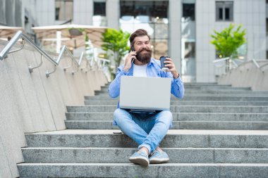 Cep telefonuyla konuşan, merdivenlerde kahve içen mutlu bir adam. Gülümseyen adam dışarıdaki bilgisayarı kullanarak cep telefonu görüşmesi yapıyor. Mobil iletişim. Telefon görüşmesi. Cep telefonu çağrısı.