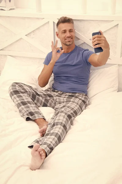 stock image blogging man with phone wear pajama, ok. blogging man with phone in bed. photo of blogging man with phone at home. blogging man with phone in bedroom.