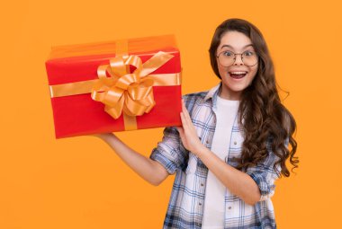Şaşkın ergen çocuk hediye kutusunu tutuyor. Sarı arka planda izole edilmiş genç bir çocuk. Gözlüklü genç bir çocuk. Stüdyodaki gençlik fotoğrafı..