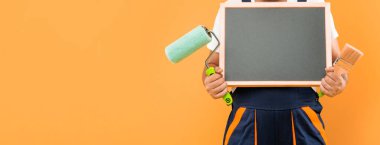 İş kıyafetleri içinde kırpılmış adam dekoratörü sarı arka planda fotokopi alanı olan boya fırçası ve karatahta tutuyor..