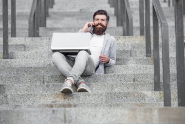 Mutlu adam cep telefonuyla konuşuyor merdivenlerde dinleniyor. Gülümseyen adam cep telefonuyla konuşuyor ve dizüstü bilgisayarla çalışıyor. Mobil internet üzerinden sesli arama yapıyorum. Mobil iletişim. Telefon görüşmesi.