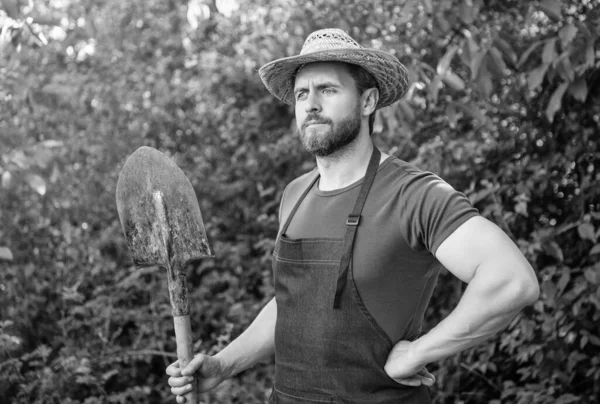 Thoughtful farmer man in farmers hat and gardening apron holding garden shovel natural outdoors.