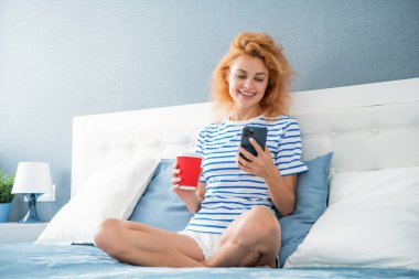 Kadın telefonda internette sörf yaparken gülümsüyor. Evde telefonuyla internette sörf yapan bir kadın. Kadının evinde internet sörfü.