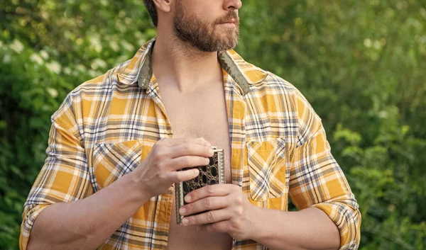 cropped view of man with whiskey flask. man with whiskey flask outdoor. man with whiskey flask wear checkered shirt. photo of man with whiskey flask.