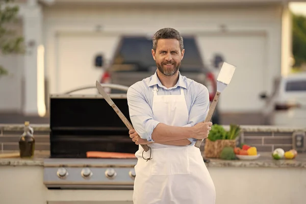 image of chef man at barbecue and grill in apron. chef man at barbecue and grill. chef man at barbecue and grill hold utensil. chef man at barbecue and grill outdoor.