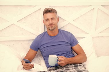 Yatakta kahve içerken cep telefonundan sabah haberlerini okuyan kır saçlı adam. Mobil cihazda mobil internet kullanılıyor. Mobil iletişim.