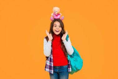 Bilgi günü. Eğitim konsepti. Sarı arka planda sıradan giyinmiş bir çocuk. 1 Eylül. Mutlu çocukluk. Oyuncak ayılı çocuk. Şok olmuş genç kız sırt çantası taşıyor. Okula geri dön..