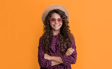 Hasır şapkalı, güneş gözlüklü, uzun, kıvırcık saçlı, sarı arka planda gülümseyen çocuk..