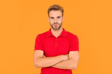 Erkek modası. Erkek güzelliği. Kırmızı tişörtlü bir adamın portresi. Genç adam sarı arka planda ellerini çapraz yaptı. Saç ve sakal bakımı. Kendine güvenen ve yakışıklı, tıraşsız adam..