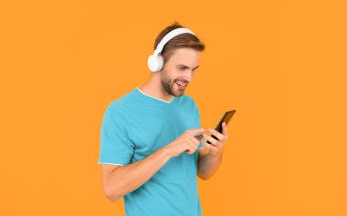 Cep telefonundan mesaj atan, kulaklıkla dijital müzik dinleyen mutlu genç adam sarı arka planda, SMS.