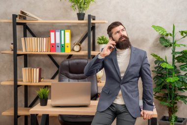 Takım elbiseli ciddi bir çalışan cep telefonuyla konuşuyor. Ofis masasında oturuyor, iletişim kuruyor..