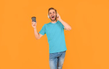 Tıraşsız, kulaklıklı, sarı arka planda kahve içen, müzik çalan genç adam..