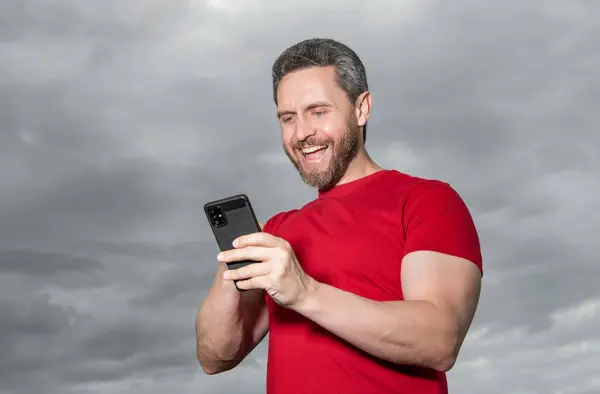 照片中的快乐男人博客类型的电话留言 男性博主在天空背景下键入电话讯息 男子博主在室外输入电话留言 男子博客类型的电话留言穿红衫 — 图库照片
