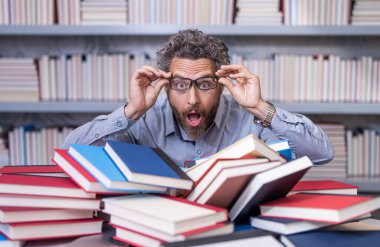 Erkek öğretmen kitap okudu. Üniversitede okul eğitimi. Bilgi günü. Kitap kurdunun fazla çalışması. Okul kütüphanesinde dahi öğretmen. Bir yığın kitabın yanında ev ödevi edebiyatı okuyor. İşini planlıyordu..