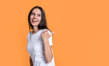 Parmakla işaret eden mutlu kız. Reklam afişi için ürün sunan kız. Sıradan tişörtlerle gülümseyen bir kız. Gülümseyen kız stüdyosu sarı arka planda izole edilmiş, kopyalama alanı.