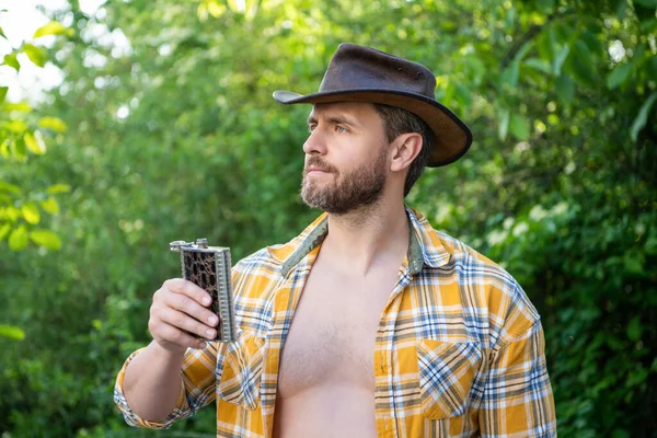 western man hold whiskey flask outdoor. western man with whiskey flask wear checkered shirt. photo of western man with whiskey flask. western man with whiskey flask.