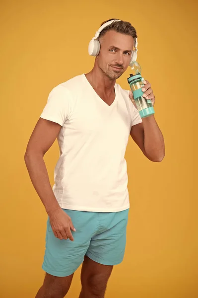 healthy sporty man drink water isolated on yellow background. sporty man drink water in studio. sporty man drink water after training. sporty man in headphones drink water.