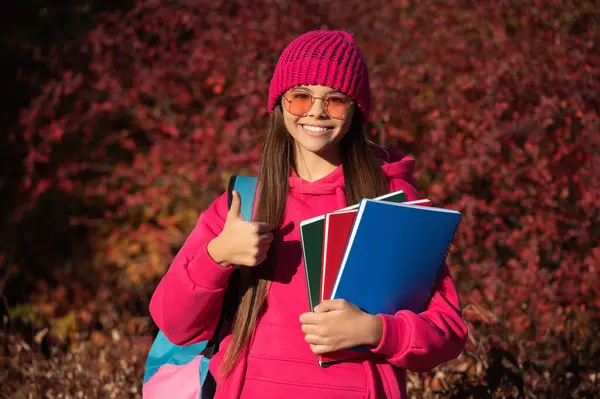 stock image knowledge and education. school knowledge. back to school. autumn girl holding school homework. knowledge for adolescence. teen girl in autumn park outdoor. autumn is a time to study. thumb up.