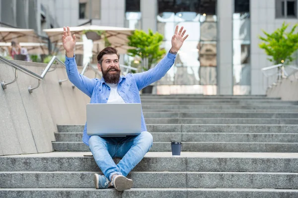 Happy freelancer gesturing hello sitting on stairs. Male freelancer working online outdoors. Freelancer man using laptop computer for remote work online. Freelancer freelancing online.