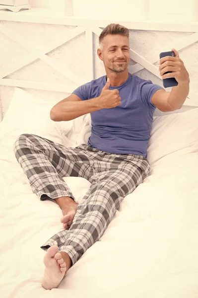 smiling video call man with phone wear pajama. video call man with phone in bed. photo of video call man with phone at home. video call man with phone in bedroom.