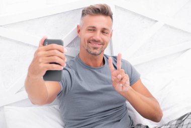 selfie man with phone in bed, peace. photo of selfie man with phone at home. selfie man with phone in bedroom. selfie man with phone wear shirt.