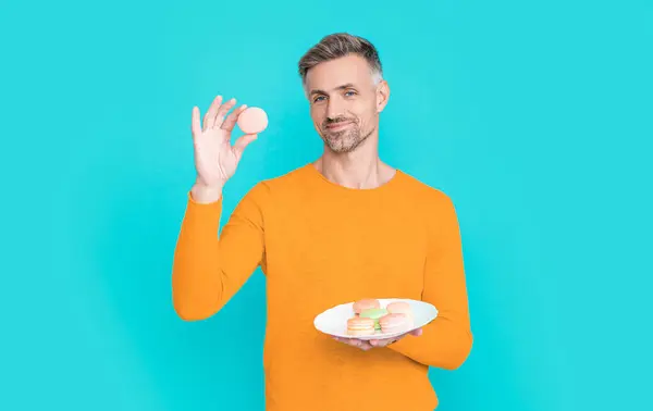 stock image photo of smiling man hold yummy macaroon. man hold yummy macaroon isolated on blue. man hold yummy macaroon in studio. man hold yummy macaroon on background.