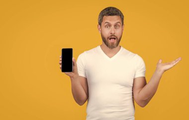 Sarı arka planda izole edilmiş telefon ekranı gösteren şaşırmış bir adam. Stüdyoda telefon ekranı var. Adam telefon ekranını gösteriyor. Adam kopyalama alanı ile telefon ekranını gösteriyor.