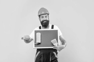 cheerful man painter in helmet and work clothes hold paint roller brush and blackboard with copy space on yellow background.