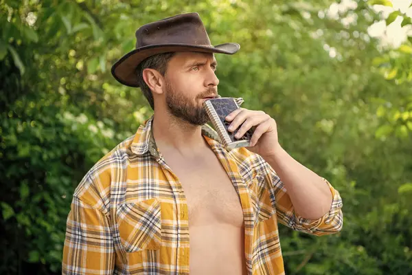 stock image masculine cowboy drink from flask wear hat. photo of masculine cowboy with flask. masculine cowboy with flask. masculine cowboy with flask outdoor.