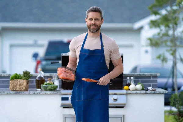 cheerful man cook salmon in apron. photo of man cook salmon food. man cook salmon on grill. man cook salmon outdoor.