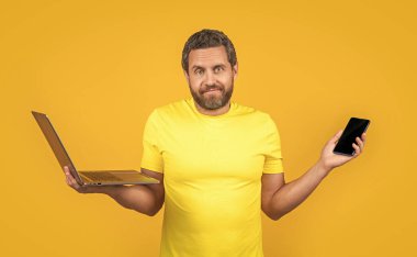 İnternette bilgisayar ve telefonla çalışan kafası karışmış bir adamın fotoğrafı. Çevrimiçi çalışan bir adam sarıda yalnız çalışıyor. Adam internette arka planda çalışıyor. Stüdyoda çalışan adam..