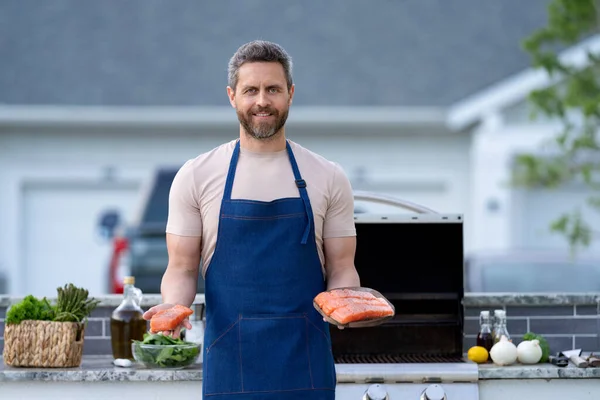 stock image glad bbq man with salmon fish. bbq man with salmon fish outdoor. bbq man with salmon fish in apron. photo of bbq man with salmon fish.