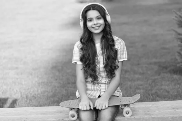 stock image photo of positive teen girl with skateboard listen music. teen girl with skateboard outdoor. teen girl with skateboard in headphones. teen girl with skateboard outside.