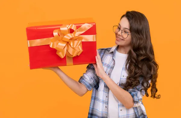 positive teen child in glasses with present. studio shot of teen child hold present. teen child holding box of present. teen child with present isolated on yellow background.