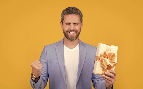 glad guy with present box for holiday. photo of guy holding occasion present box. guy hold present box isolated on yellow background. guy hold present box in studio.