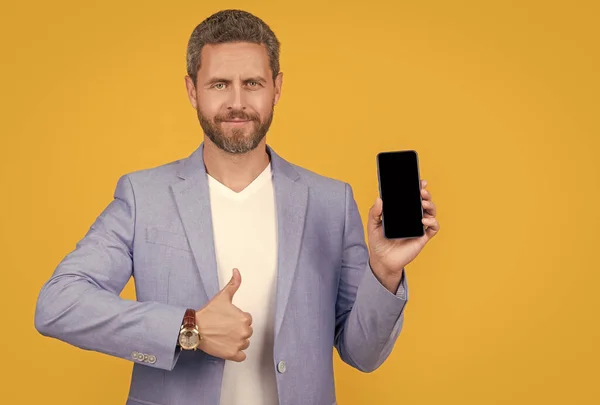 businessman showing portable phone, thumb up. businessman presenting screen of portable phone with copy space. businessman with portable phone isolated on yellow. businessman hold portable phone