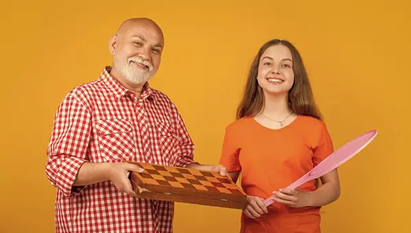 stock image smiling teen child with grandfather with badminton racket and chess on yellow background. fathers day