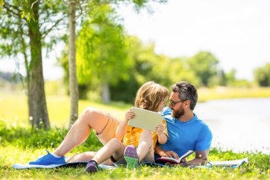 Baba oğul eğitimini kitap ve tabletle şekillendirir. Baba ve oğul parkta rahatlayın. çocukluk okulu eğitimi. Oğlu ve babası yazın kaynaşıyorlar. Baba ve oğul eğitimi. Açık havayı keşfet.