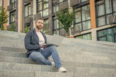 Sokakta oturan serbest çalışan bir adam. Dışarıda polisle serbest çalışan bir adam. Bilgisayarlı serbest çalışan adamın fotoğrafı. Dışarıda dizüstü bilgisayarı olan serbest çalışan bir adam..